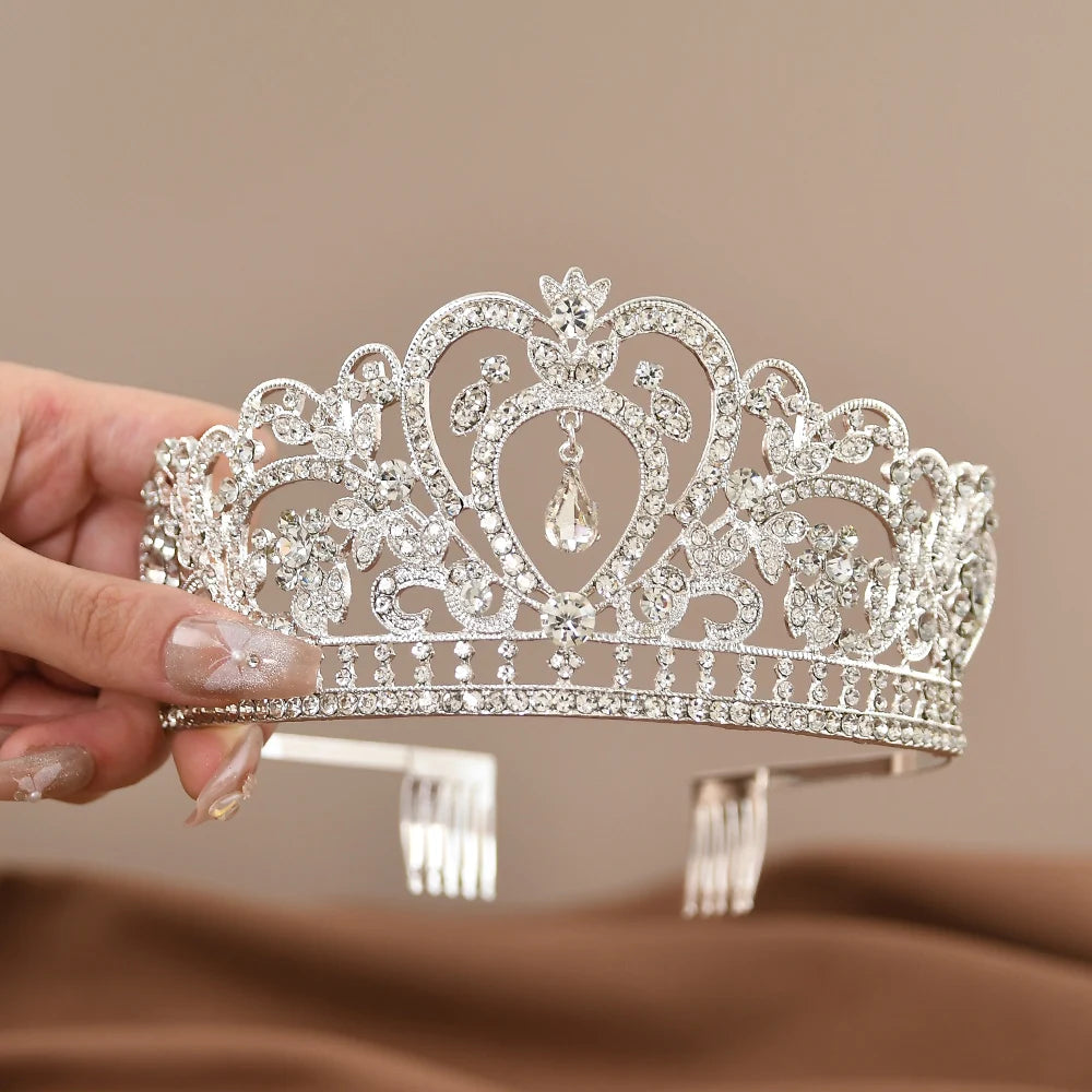 A hand holding an ornate silver tiara adorned with sparkling rhinestones and floral patterns. The tiara features a central teardrop-shaped gem, set against a soft brown background.