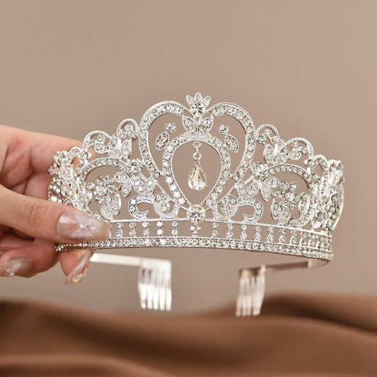 A hand holding an ornate silver tiara adorned with sparkling rhinestones and floral patterns. The tiara features a central teardrop-shaped gem, set against a soft brown background.