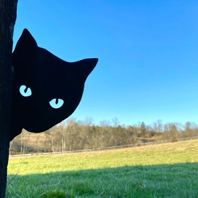 Silhouette of a cat peeking from behind a tree against a clear blue sky and a grassy field in the background.