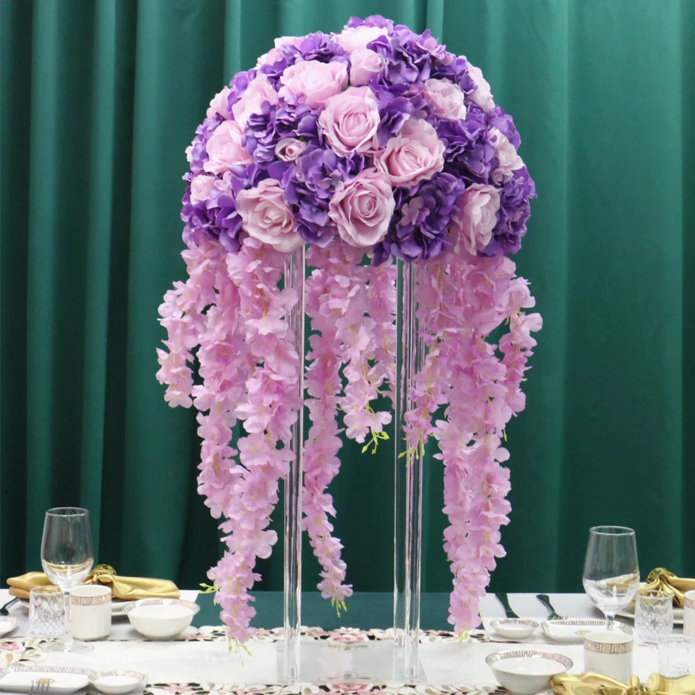 Elegant floral centerpiece with a dome of pink and purple roses, accented by cascading pink flowers, set on a table with glassware and a dark green backdrop.