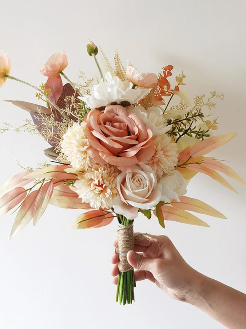 A hand holds a delicate bouquet of peach and white flowers, including roses and peonies, accented with pale green leaves and wrapped in twine against a plain background.