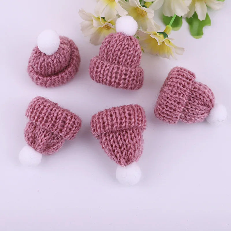 Five small pink knitted hats with white pom-poms are arranged on a surface with delicate white flowers in the background.