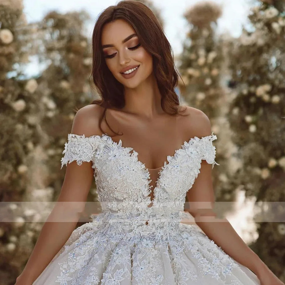 Woman in a floral lace wedding dress stands smiling, surrounded by blurred floral background. Her gown features off-the-shoulder sleeves and intricate embroidery.