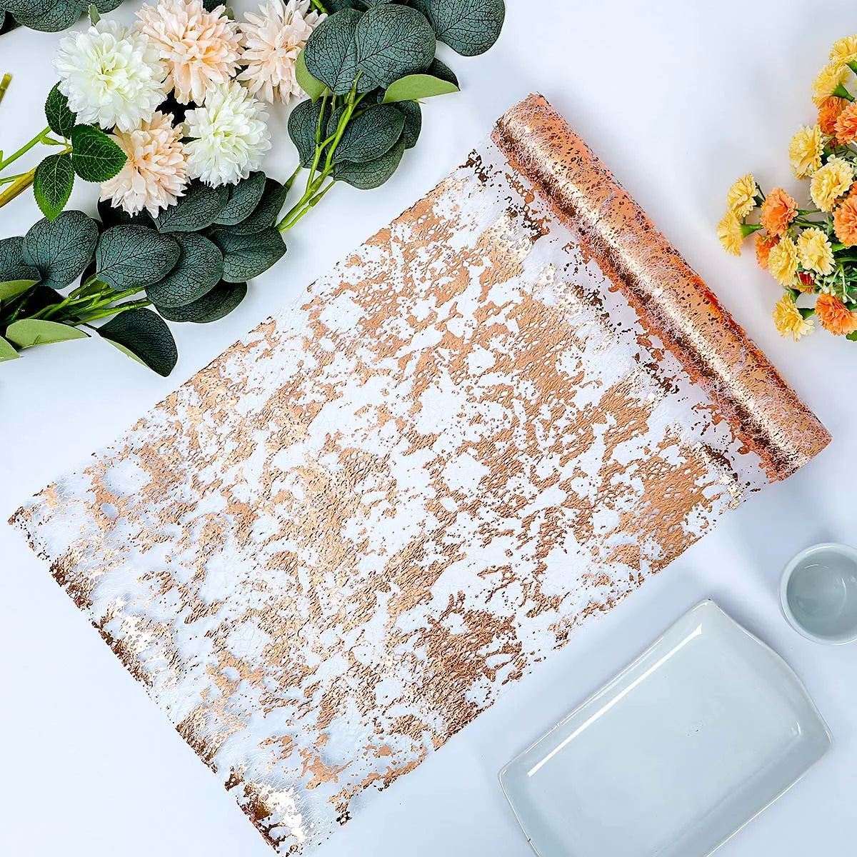 Elegant table setting with a rolled metallic rose gold and white decorative runner, flanked by cream and orange flowers on a white surface with dishware.