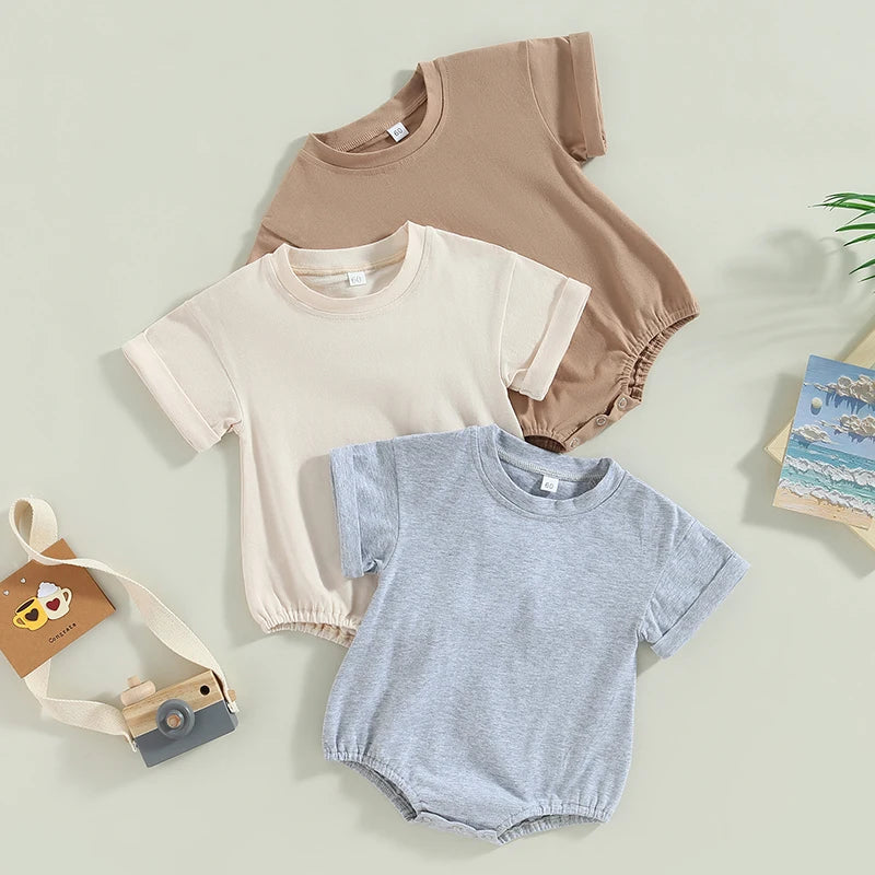 Three baby rompers in brown, cream, and gray are laid out on a soft green background. A wooden toy camera with a beige strap is nearby, along with a postcard featuring a beach scene.