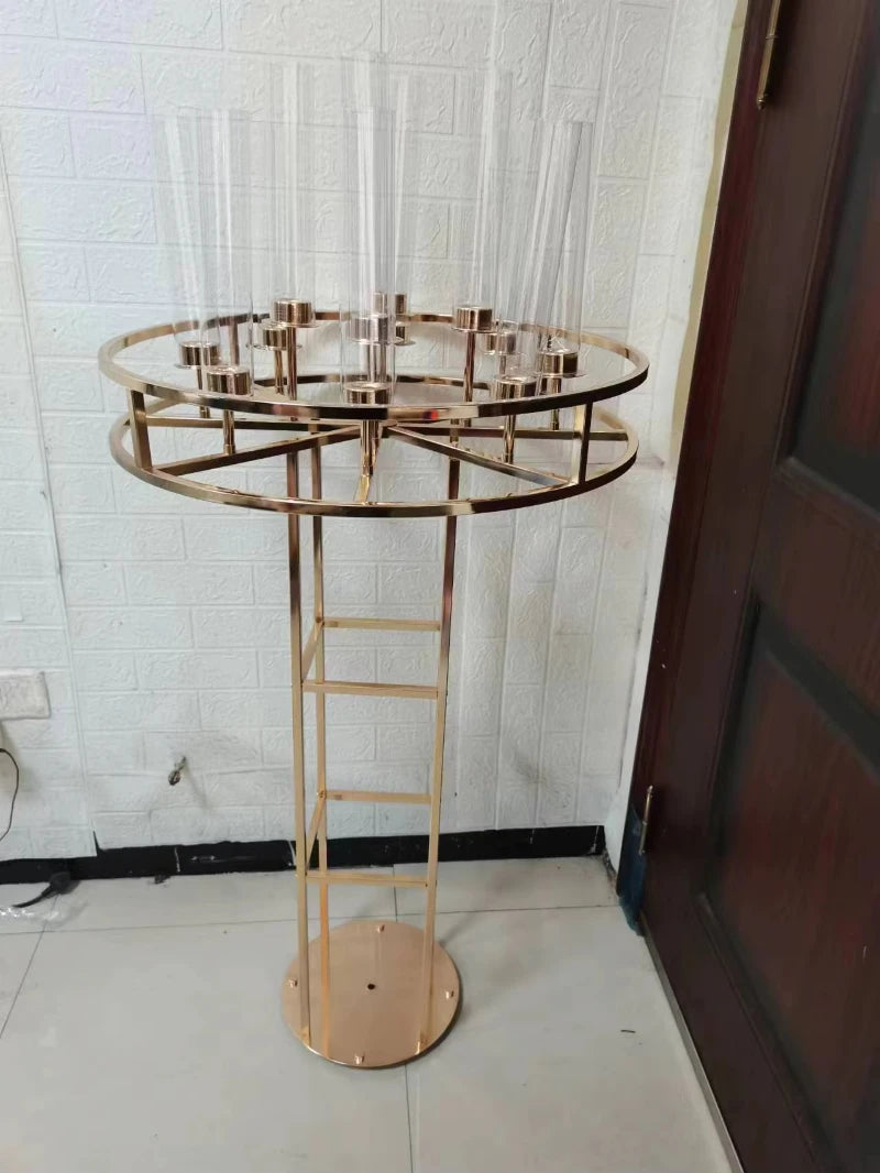 A tall, gold metal stand with a round base supports a circular frame holding multiple transparent glass cylinders against a textured white wall and dark wooden door.