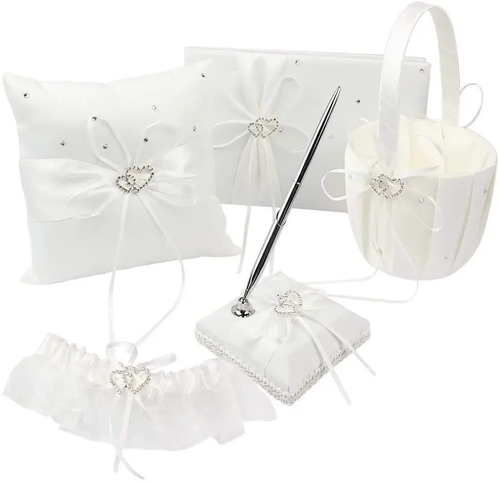 A collection of wedding accessories including a pillow, guest book, and basket, all adorned with white satin ribbons and heart-shaped rhinestone embellishments.