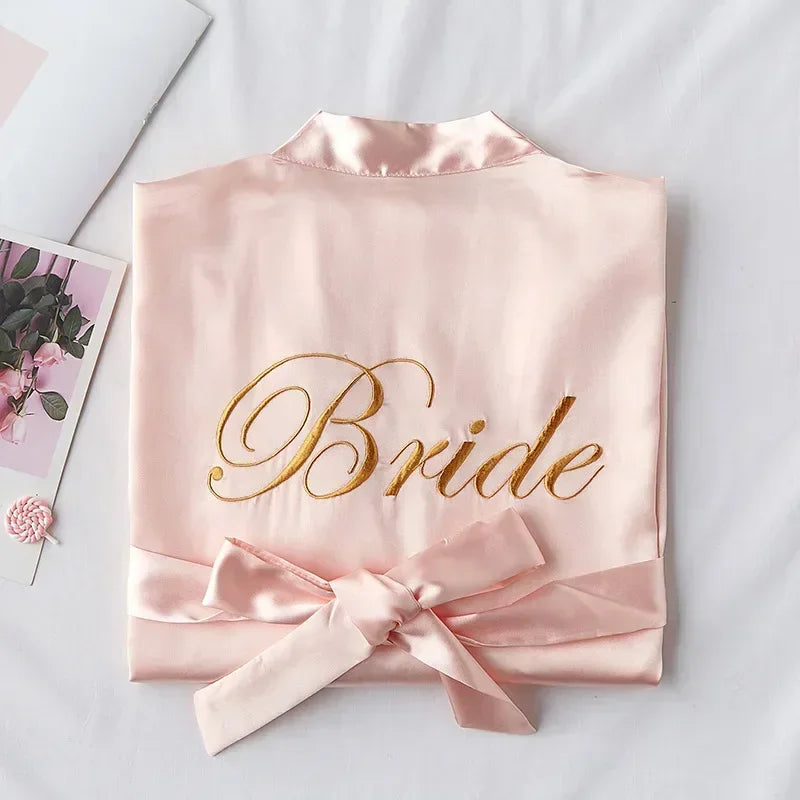 Pink satin robe with "Bride" embroidered in gold script, neatly folded with a bow. A floral photo and crochet flower lie nearby on a white surface.