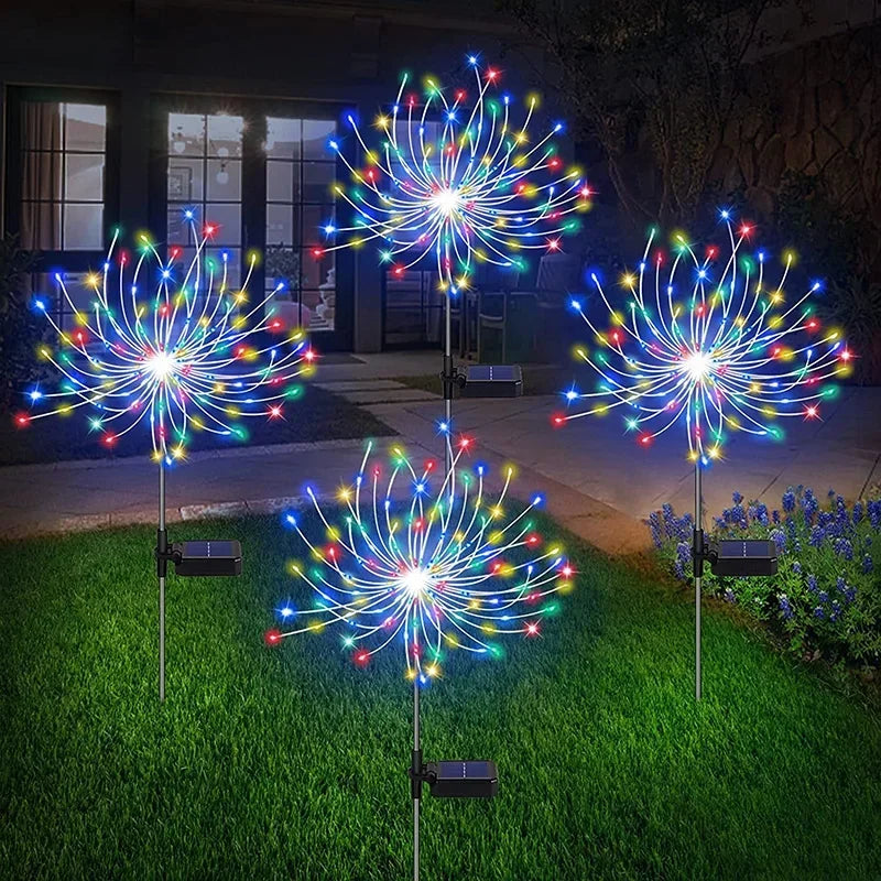 Four colorful LED garden lights resembling fireworks illuminate a lawn at night. Each light features solar panels, creating festive bursts of blue, red, green, and yellow.