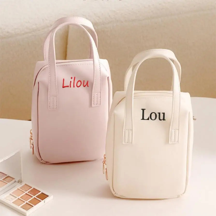 Two small handbags with short handles, one pink labeled "Lilou" and one cream labeled "Lou," sit on a table beside a small open eyeshadow palette.