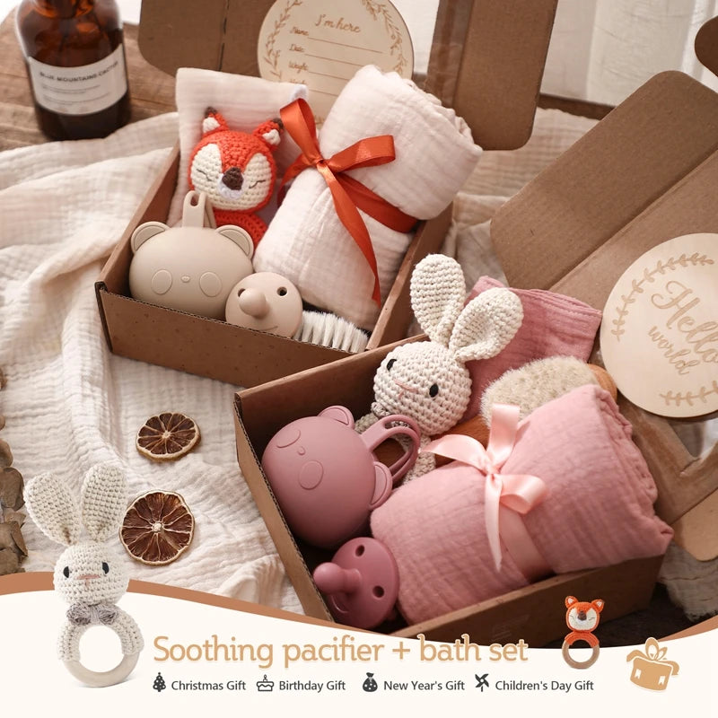 Gift box with baby bath and pacifier set. Includes knit toys shaped like animals, pink and beige blankets tied with ribbons, wooden decorations, and dried orange slices.