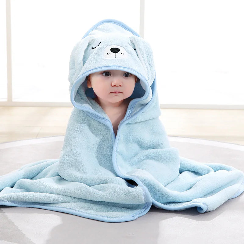 A baby in a blue, hooded animal-themed blanket sits on a light gray circular rug. The hood features a cute bear face with floppy ears, creating a cozy and playful look.