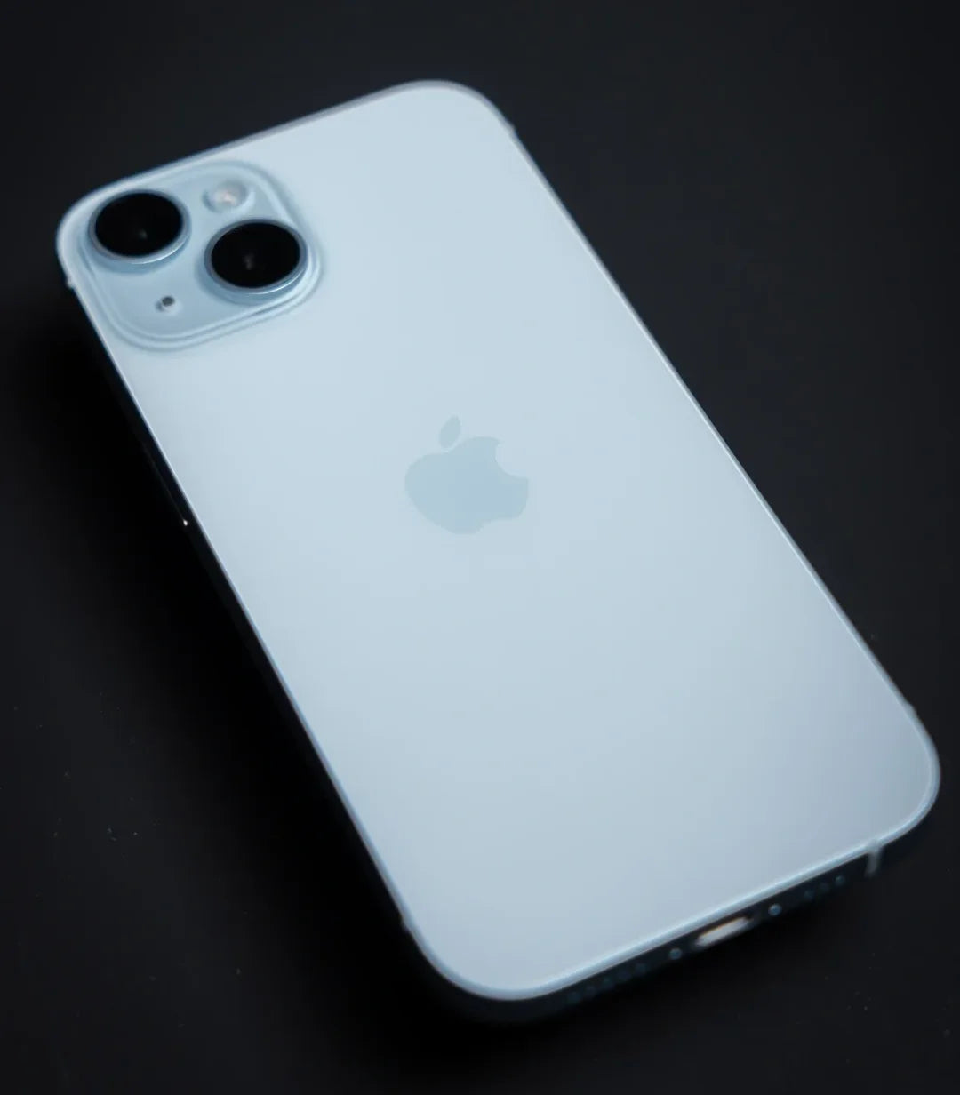 Close-up of a light blue smartphone on a dark background, featuring dual rear cameras and an Apple logo in the center.
