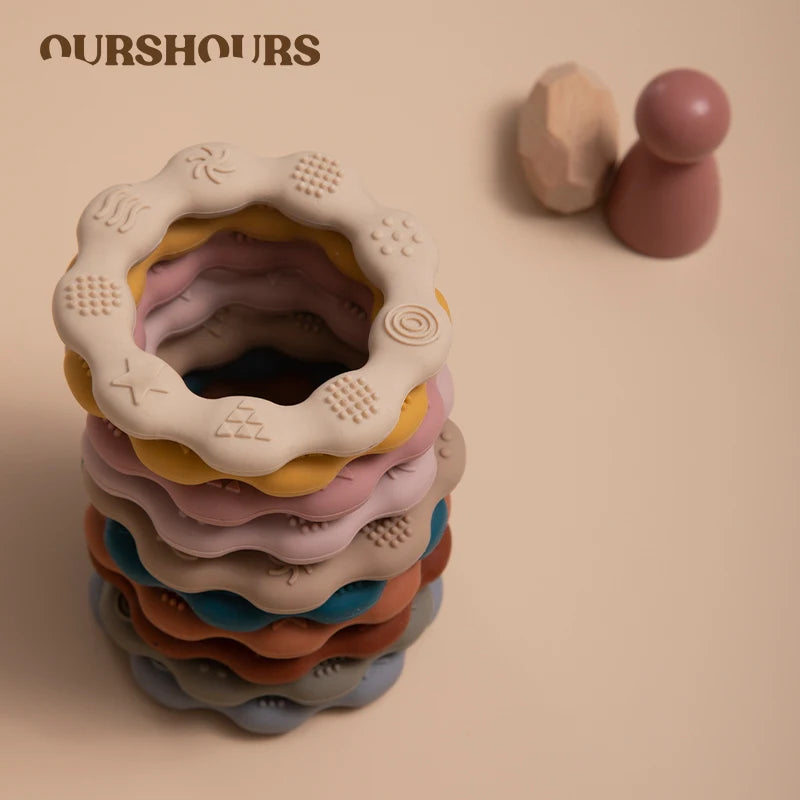 A stack of colorful textured rings with embossed patterns is shown, accompanied by a wooden peg toy in the background. The image has a soft beige background and "OURSHOURS" text.