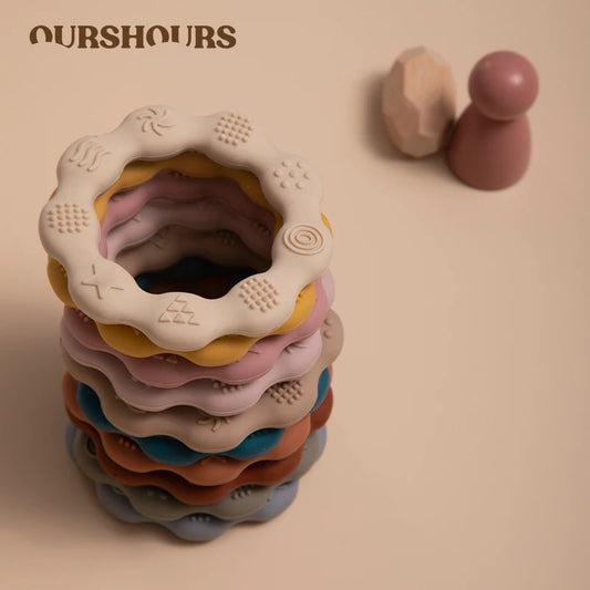 A stack of colorful textured rings with embossed patterns is shown, accompanied by a wooden peg toy in the background. The image has a soft beige background and "OURSHOURS" text.