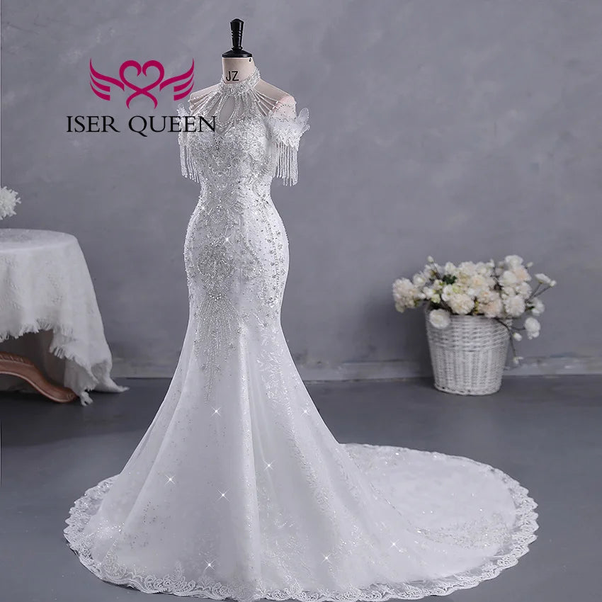 Elegant white wedding dress on a mannequin with lace details and beaded fringe sleeves. A long train flows from the fitted silhouette, with a basket of white flowers nearby.