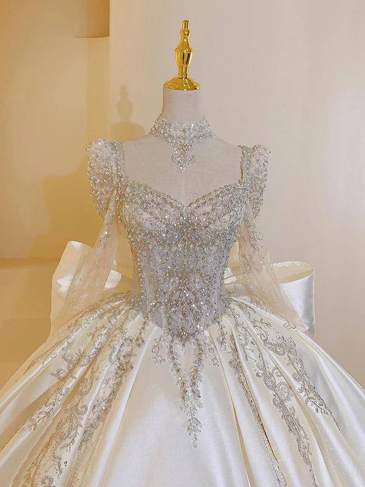 Elegant white gown with intricate silver embroidery and beadwork on the bodice and sleeves, featuring a high collar and a large satin bow, displayed on a mannequin.