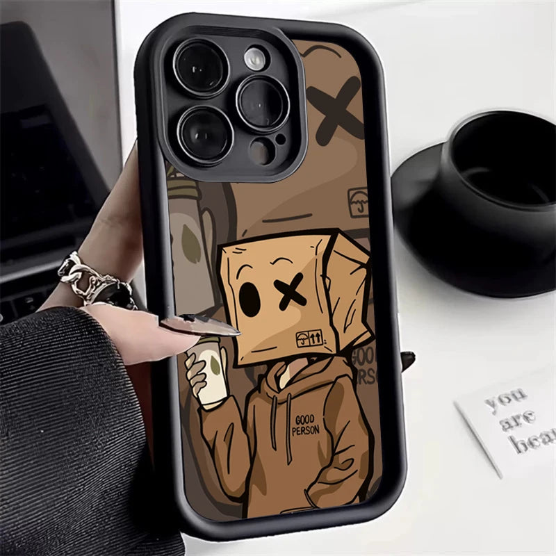 iPhone with cartoon case featuring a hooded figure with a box head holding a drink. The box has a cross for one eye, and the hoodie reads "Good Person." Nearby is a black cup.