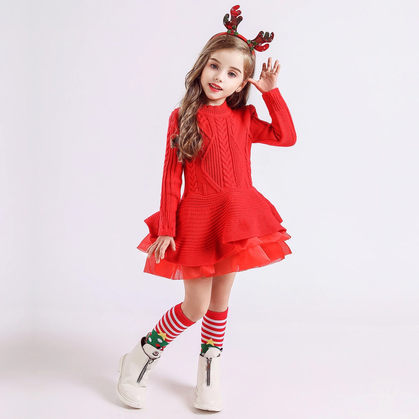 Young girl in a red knitted dress and reindeer antler headband, posing joyfully. She wears Christmas-themed striped socks and white boots, standing against a plain background.