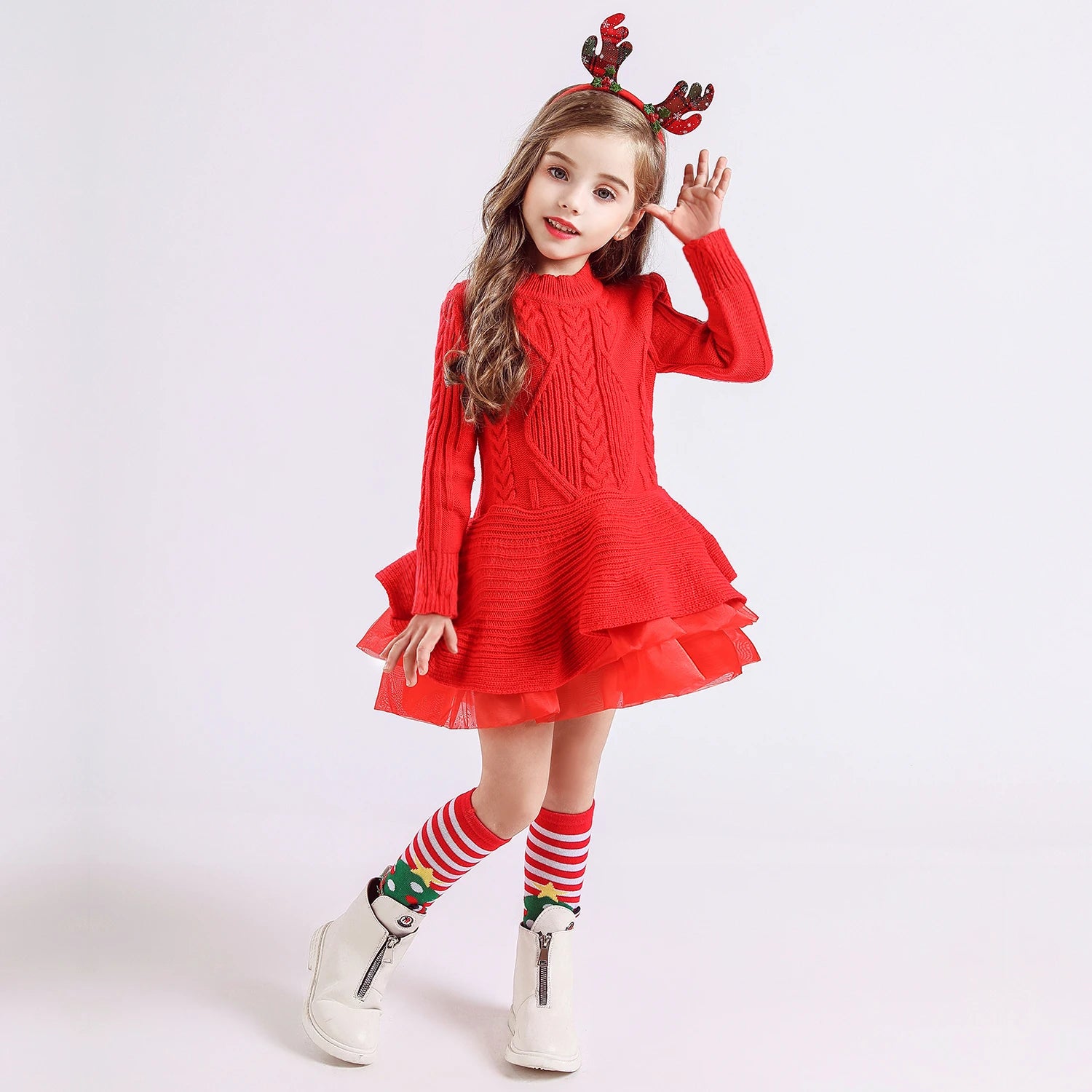 Young girl in a red knitted dress and reindeer antler headband, posing joyfully. She wears Christmas-themed striped socks and white boots, standing against a plain background.