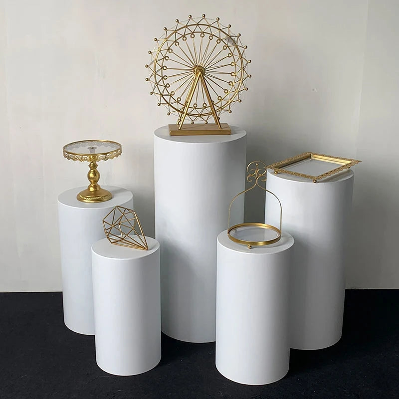 Five white cylindrical pedestals display gold decor: a Ferris wheel, tray, geometric sculpture, round frame, and square frame, set against a plain background.