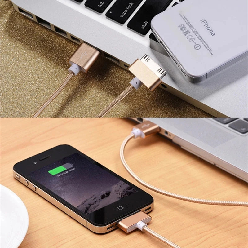 Charging cables connecting an older iPhone to a laptop. The phone displays a charging icon, and the beige braided cables are plugged into the laptop's USB ports.