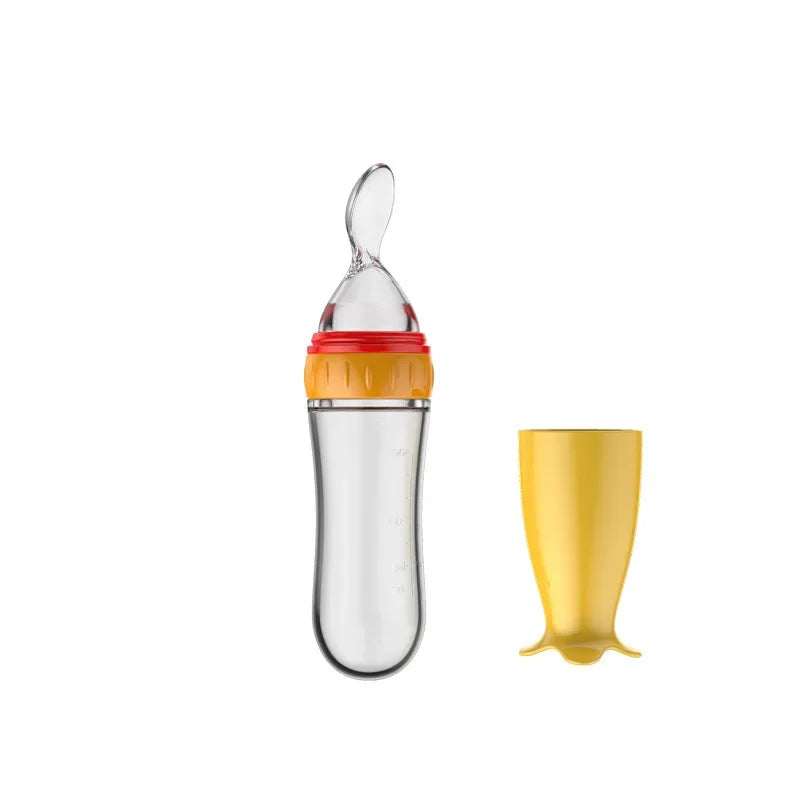 Clear baby bottle with red and orange accents, featuring a built-in spoon attachment. Includes a yellow cap beside it. Designed for easy feeding.