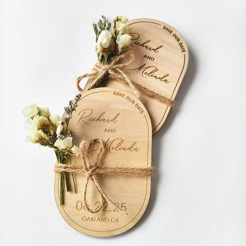 Wooden save-the-date magnets feature names "Richard and Melinda," a date of 06.22.25, and location "Oakland, CA." Tied with twine and decorated with small white flowers.