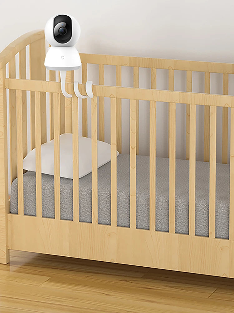 A light wood crib with a gray mattress and a white pillow. A white baby monitor camera is mounted on the crib rail, positioned to view the interior of the crib.