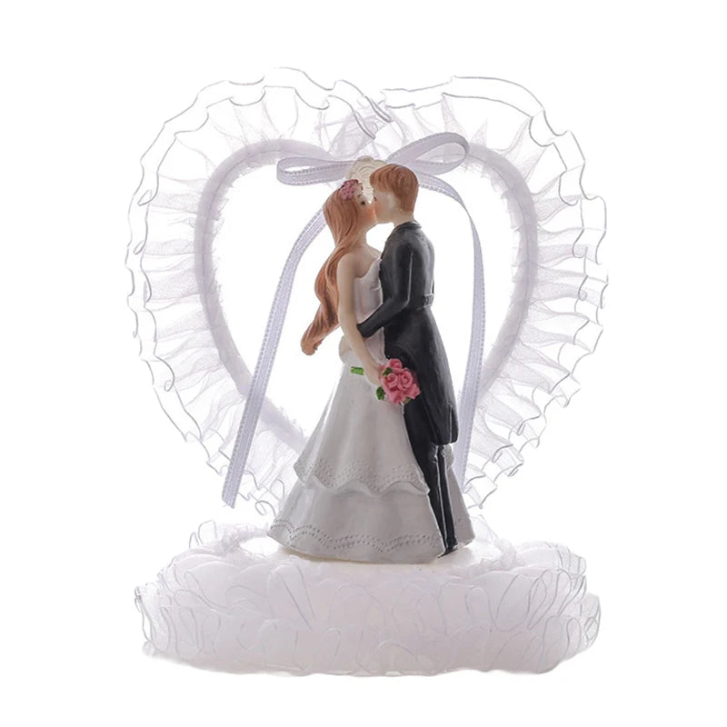 Bride and groom figurine kissing, with the bride in a white dress holding pink roses, and the groom in a black tuxedo. A white ruffled heart backdrop completes the decoration.