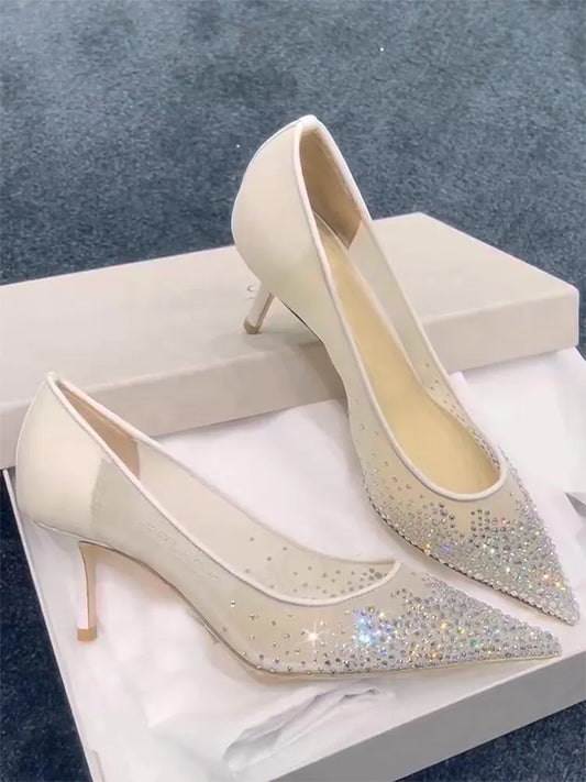 Elegant white high heels with sparkling rhinestone embellishments on pointed toes, resting on white tissue paper inside a shoebox against a textured gray background.