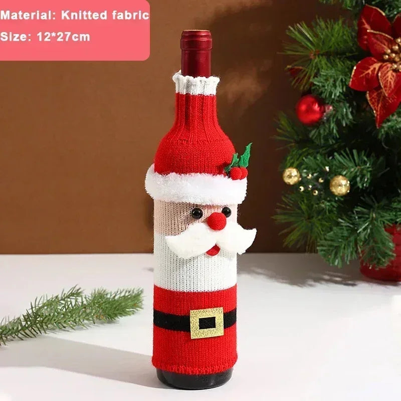 Wine bottle dressed in a Santa-themed red and white knitted cover with a belt and face details. It's placed on a white surface beside festive greenery and decorations.