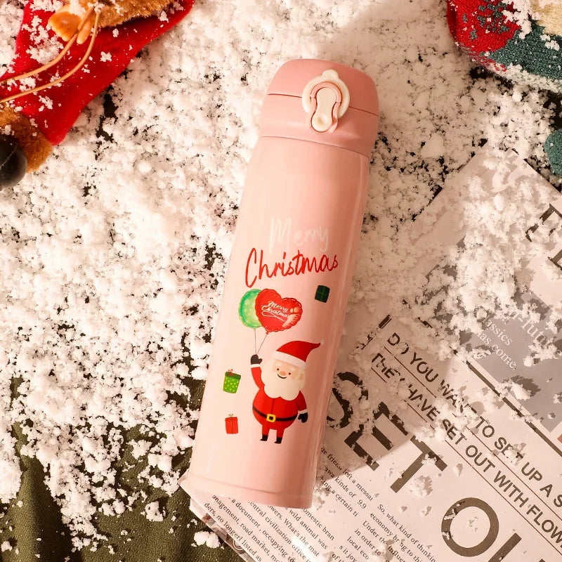 Pink thermos with Santa and "Merry Christmas" text, surrounded by faux snow, holiday decorations, and a partially visible magazine page in the snowy setting.