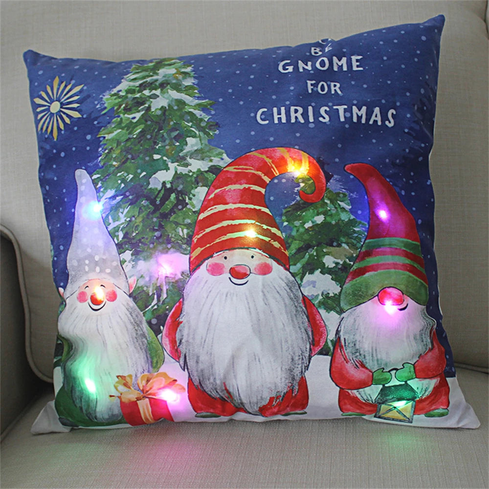 Festive pillow with lit-up gnomes in red and green hats, set against a snowy background with trees. Text reads “Be Gnome for Christmas.” Multicolored lights accent the design.