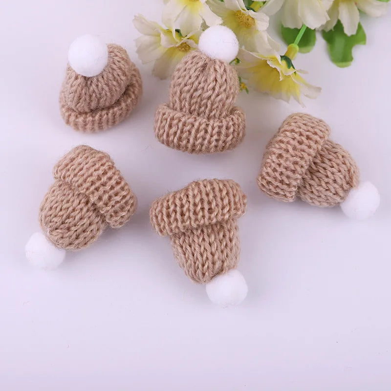 Five small, beige knitted hats with white pom-poms are scattered on a white surface, with pale yellow flowers in the background.