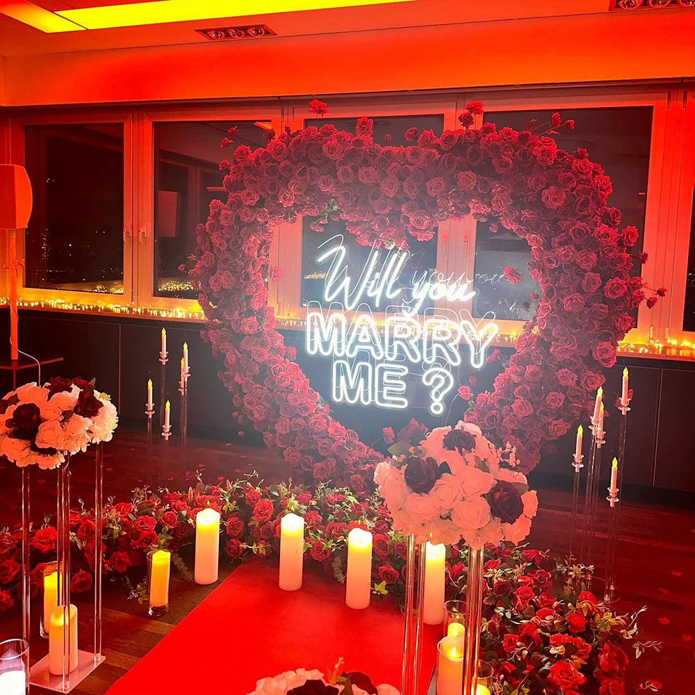 Heart-shaped rose arrangement with "Will you marry me?" in neon lights. Surrounded by candles and more flowers in a dim, romantic setting.