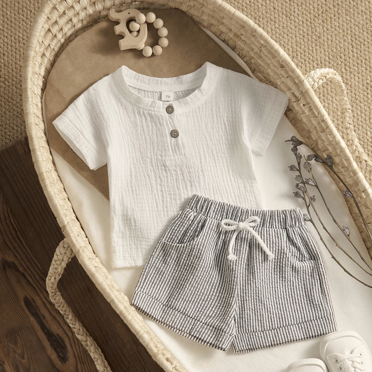 Baby outfit in a basket: white textured top with button detail, striped drawstring shorts, wooden elephant teether, and dried flowers on a neutral background.