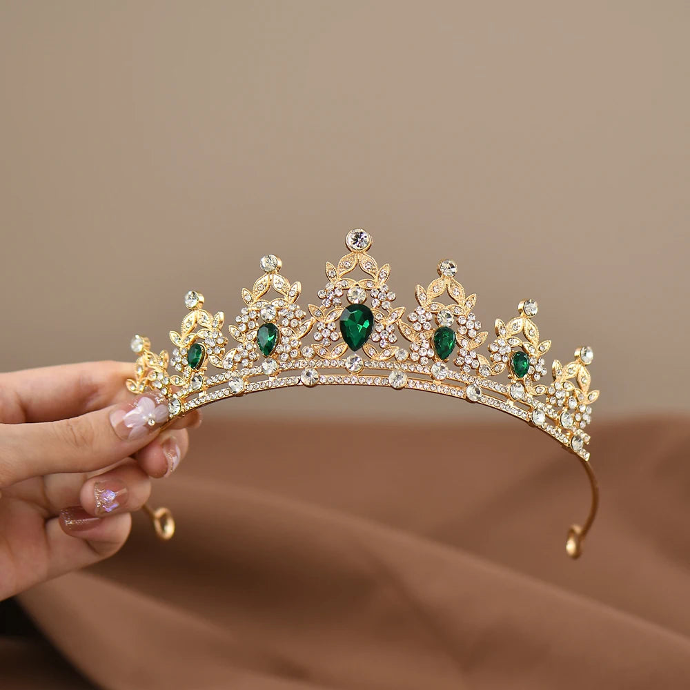 Elegant gold tiara with green gems and clear crystals held by a hand with decorated nails, set against a soft brown background.