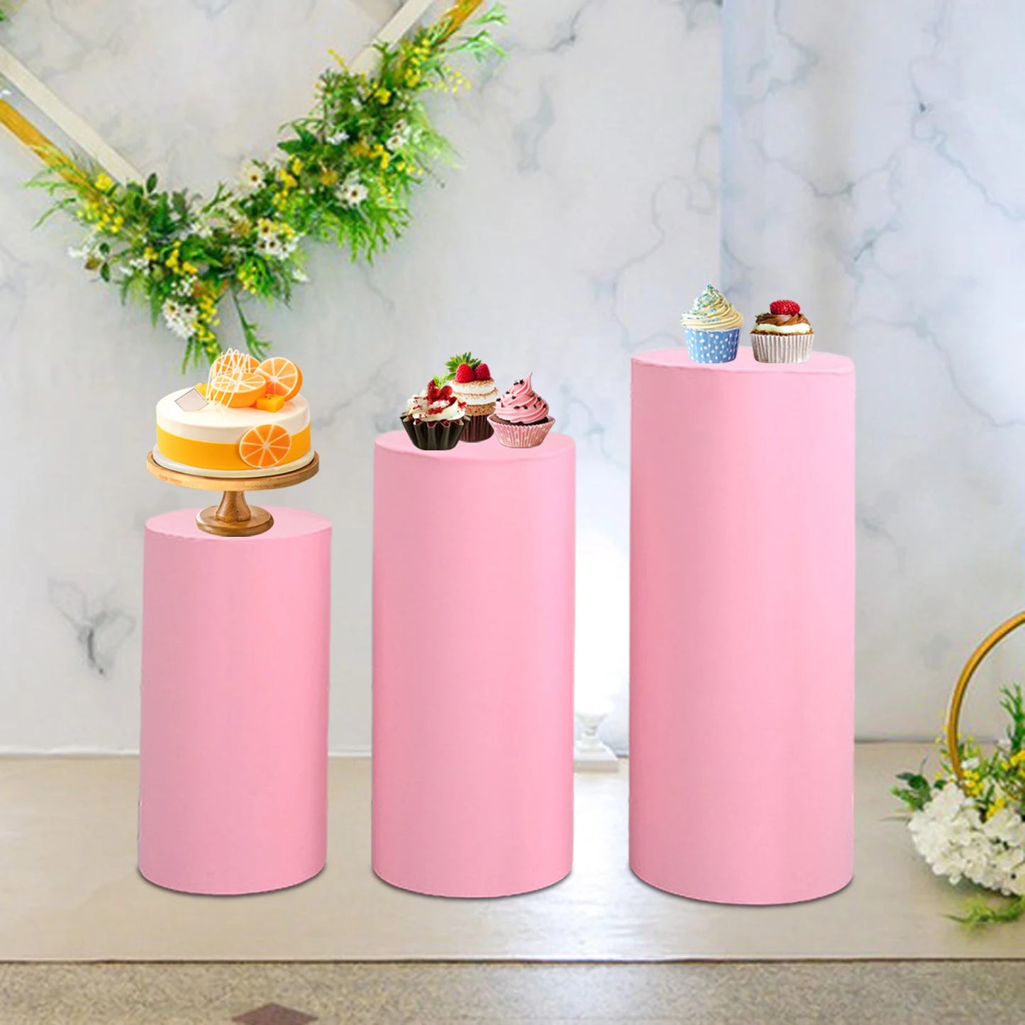 Three pink cylindrical stands display various desserts: an orange cake on the left, assorted cupcakes in the center, and additional cupcakes on the right. Floral garland in the background.