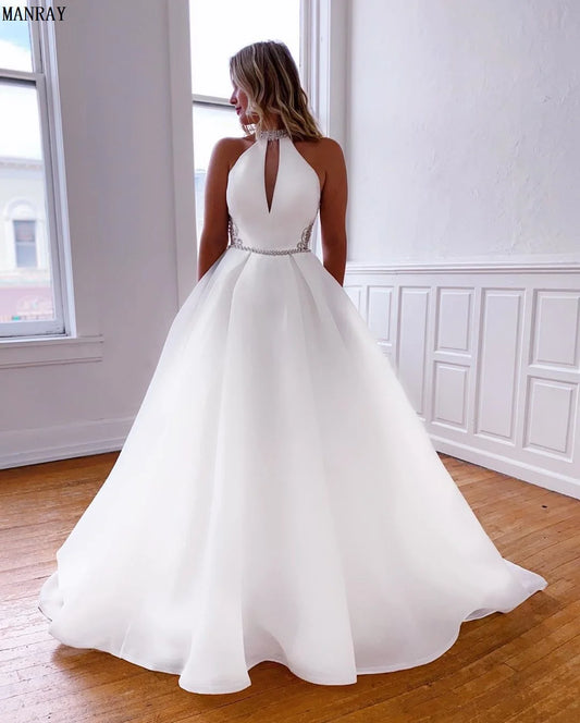 A woman in a white halter-neck ball gown with beaded details stands in a bright room with wooden floors and white paneled walls, looking out a window.