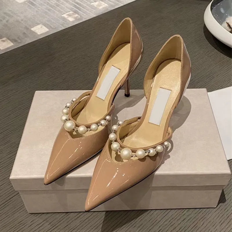 Beige patent leather high heels with pointed toes adorned with pearl beads, placed on a beige shoebox, resting on a wooden surface.