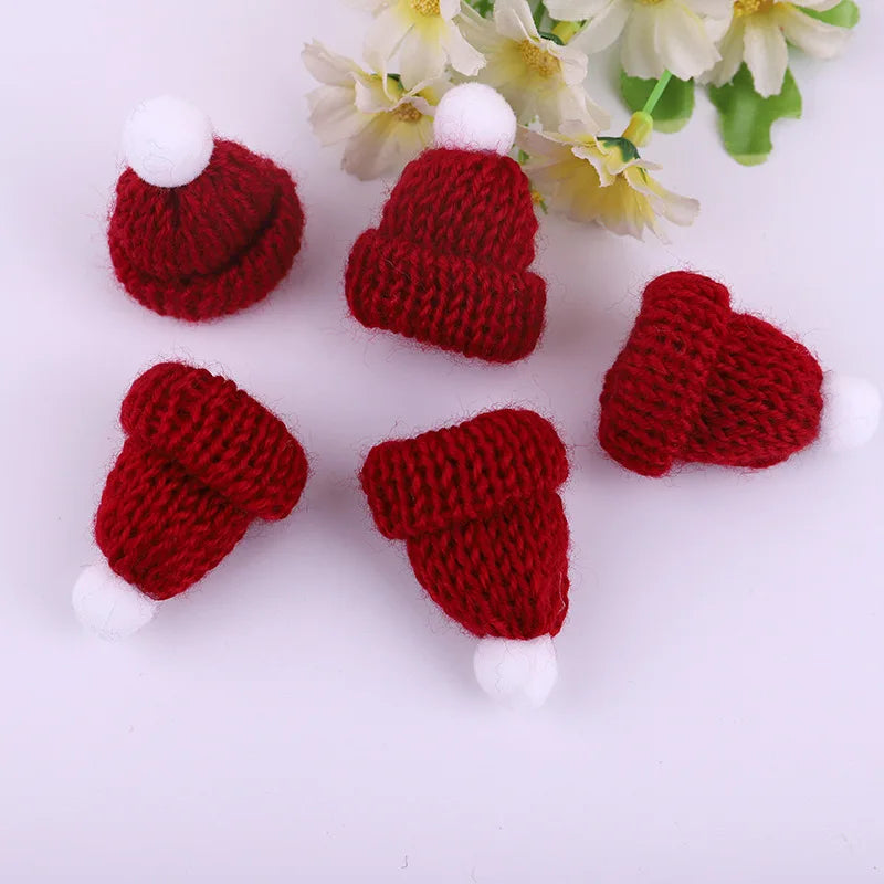 Five small red knitted hats with white pom-poms are arranged on a surface, accompanied by yellow and white flowers in the background.