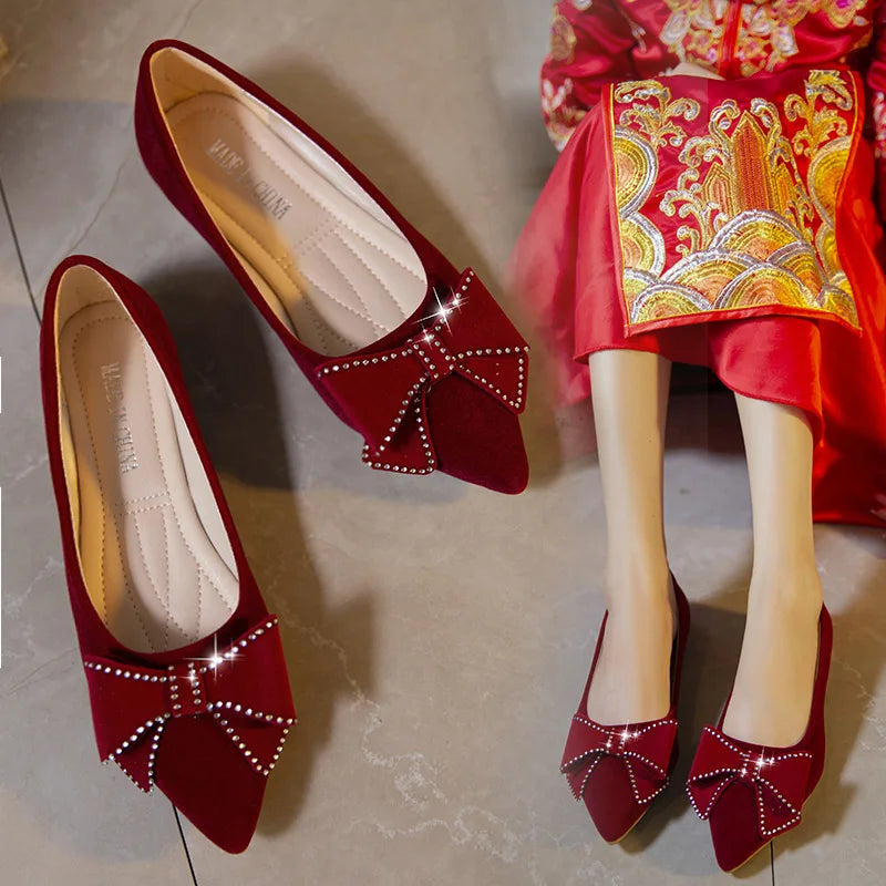Elegant red velvet flats with rhinestone-studded bows, paired with a bright red embroidered dress. The shoes glisten under the light on a textured floor.