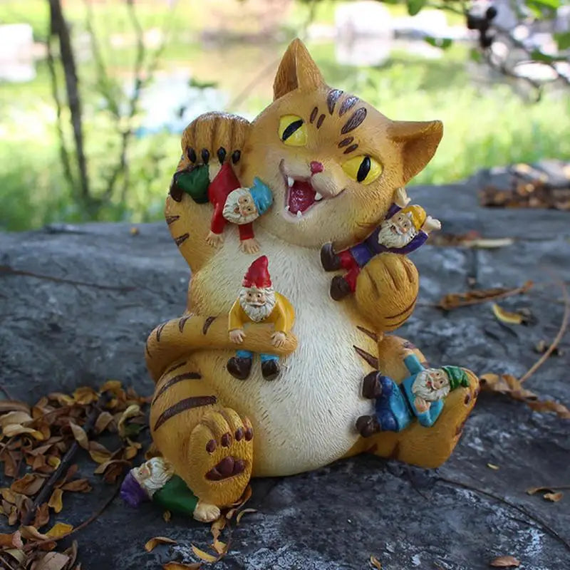 A playful garden statue of a chubby brown-striped cat on a rock, holding colorful, tiny gnomes in its paws and mouth, with a lush green background.