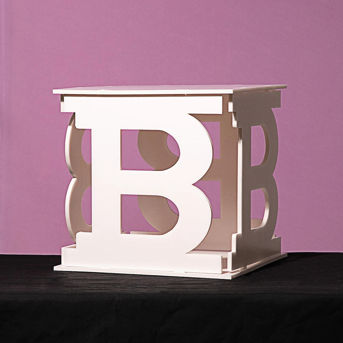 A white cube-shaped side table featuring large, cut-out "B" letters on each side. It rests on a dark surface against a pastel purple background.