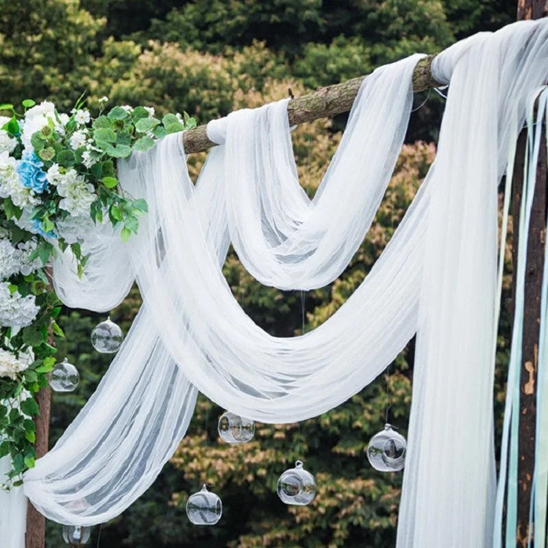 Outdoor wedding arch draped with flowing white fabric, adorned with clusters of white and blue flowers. Clear glass orbs hang beneath, set against lush green foliage backdrop.