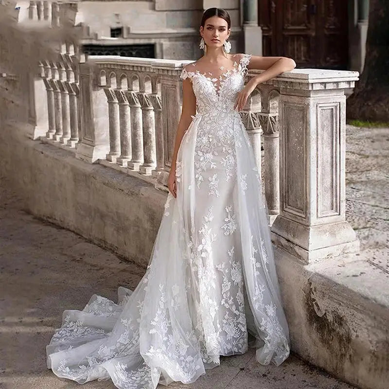 Woman in an elegant lace wedding gown with floral patterns, standing by a stone balustrade outdoors. The dress has an off-shoulder design and a flowing train.