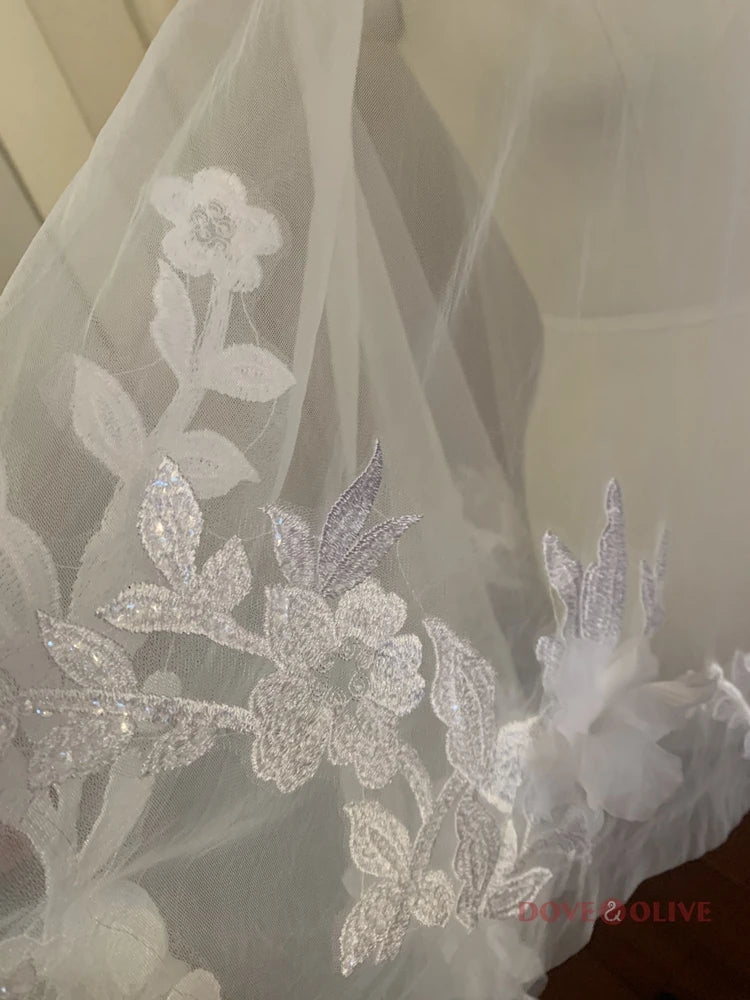 Close-up of delicate white lace fabric featuring intricate floral and leaf embroidery. The sheer material allows light to pass through, highlighting its elegant design.