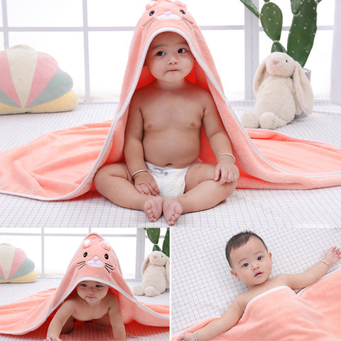 A baby in a white diaper is wrapped in a cute, orange, animal-themed hooded towel. The background includes a stuffed rabbit, a pillow, and a cactus plant.