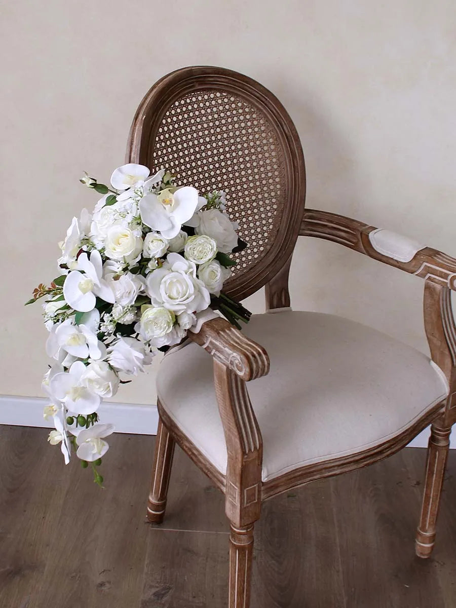 A vintage wooden chair with a cane backrest holds a cascading bouquet of white roses and orchids. The chair sits on a wooden floor against a pale wall.